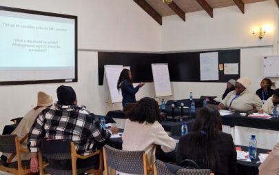 In a room with walls that have a screen displaying a PowerPoint presentation and blackboards with notes, there is a group of people, one of whom is standing in front of flipcharts while the others are sitting at tables.