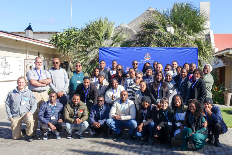 Eine Gruppe von Personen vor einem blauen Banner mit Aufschrift "Republic of Namibia" in einem Hof mit Palmen, umringt von Gebäuden.