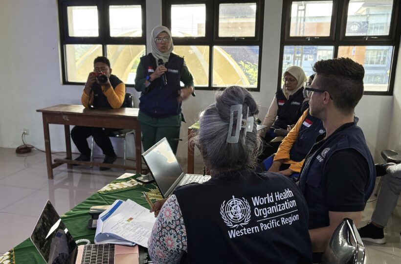 Indoors with a window front, where a small group of people is seated and a woman holding a microphone is standing. One person is wearing a vest with the inscription on the back: World Health Organization - Western Pacific Region.