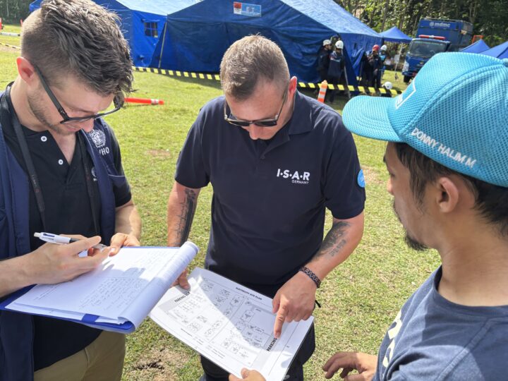 On a lawn with blue tents in the background, three men are standing in a circle and seem to discuss two documents which two of them are holding in their hands.