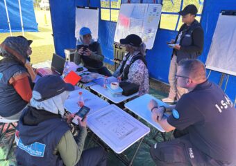 In a pavilion on a lawn, five people are sitting around a table, and another person is standing in front of a flipchart.