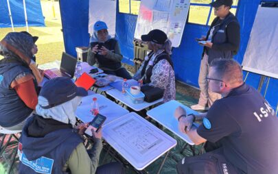 In einen Pavillon auf einer Wiese sitzen Personen um einen Tisch herum fünf Personen, eine weitere steht vor einer Flipchart.