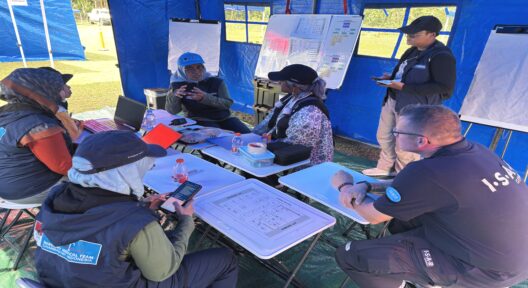 In a pavilion on a lawn, five people are sitting around a table, and another person is standing in front of a flipchart.