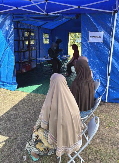 Three people wearing skirts and long scarves are sitting in a row in front of a blue tent, at the entrance of which a paper reads Pharmacy.