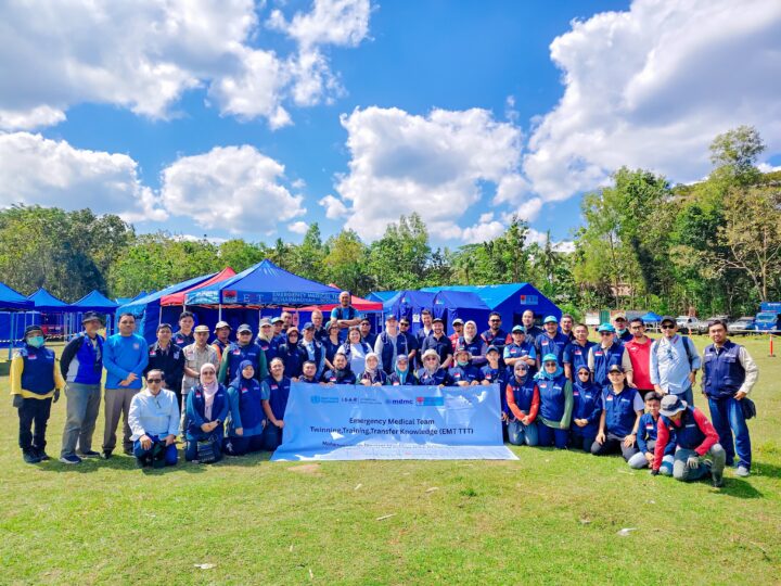 Group photo with people posing in two rows on a lawn, with blue tents, pavilions, and trees visible in the background. The front row of people is holding a banner with the title Emergency Medical Team - Twinning, Training, Transfer Knowledge (EMT TTT).
