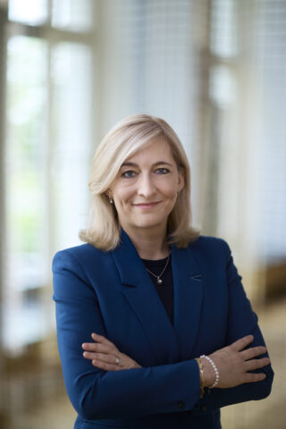 In the foreground is Federal Minister Nina Warken in a blue blazer with her arms crossed.