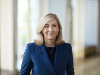 Im Vordergrund steht Bundesministerin Nina Warken in einem blauen Blazer mit verschränkten Armen.