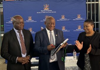Three people stand in front of the banner of the Namibian Ministry of Health. One with the PHEOC manual in his hand, the other two clapping their hands