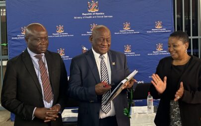 Three people stand in front of the banner of the Namibian Ministry of Health. One with the PHEOC manual in his hand, the other two clapping their hands