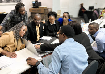 A group of eight people sitting opposite each other at a long seminar-table, all looking at the screen of a Laptop that is placed on the table.