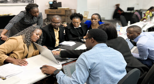 A group of eight people sitting opposite each other at a long seminar-table, all looking at the screen of a Laptop that is placed on the table.