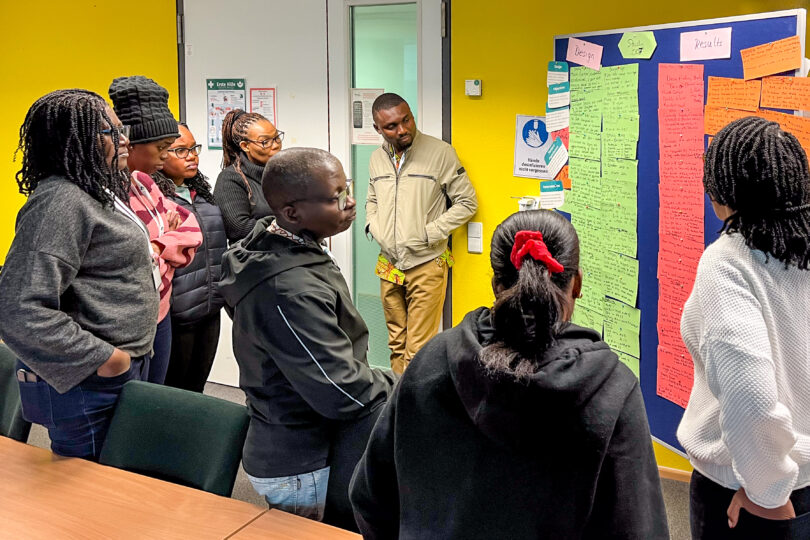 In a room: Eight people standing together looking at a pinboard deeply in thoughts . At the pinboard attached are moderation cards in various coloures carrying notes.