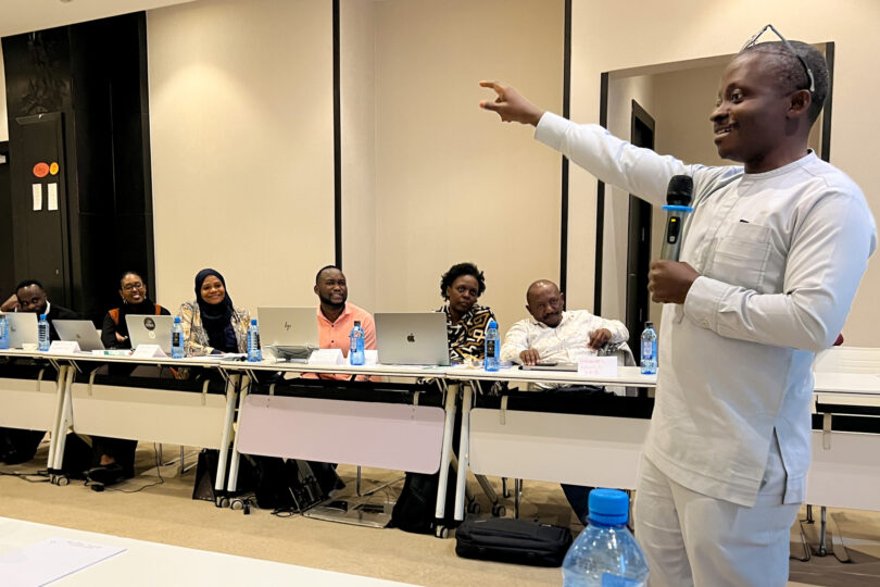 A man is standing in a room, smiling pointing at something with one hand which is raised and holding a microphone in the other hand. In the background, people are seated at a long seminar table. On the seminar table, there are laptops and water bottles.