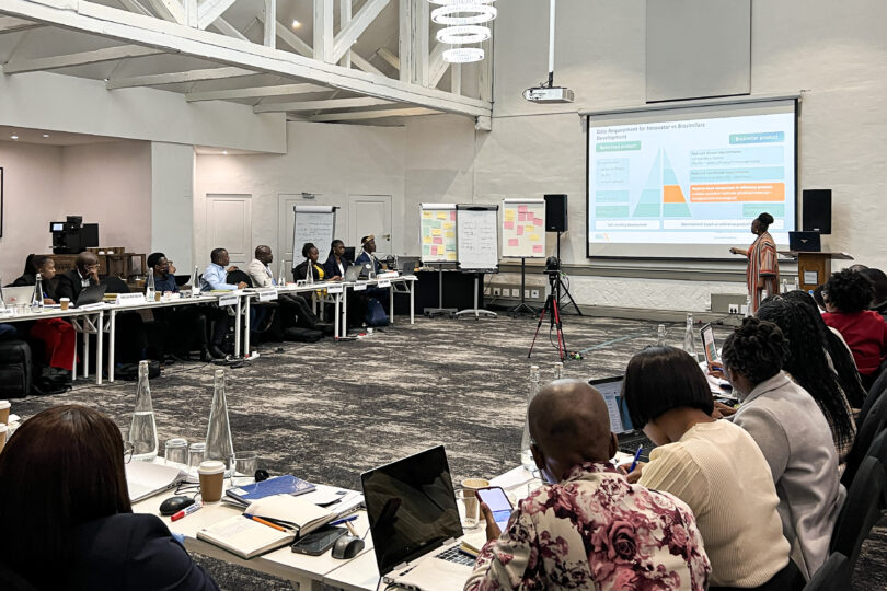 In a bigger seminar-room with u-form seminartables. People with laptops are seated at these tables. They look to the front where a woman stands in front of a big screen which shows a powerpoint presentation. From the ceiling there hangs a beamer. In addition, there are four flipcharts with notes as well as a set of speakers at the front framing the big screen.