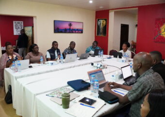 Eine Gruppe von Menschen sitzt u-förmig an zusammengeschobenen Konferenztischen mit weißen Tischdecken. Die meisten der Personen haben einen aufgeklappten Laptop vor sich auf dem Tisch und Wasserflaschen. Sie scheinen im Gespräch zu sein.