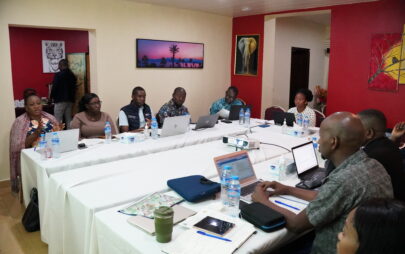 Eine Gruppe von Menschen sitzt u-förmig an zusammengeschobenen Konferenztischen mit weißen Tischdecken. Die meisten der Personen haben einen aufgeklappten Laptop vor sich auf dem Tisch und Wasserflaschen. Sie scheinen im Gespräch zu sein.