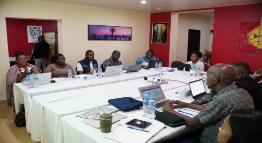 Eine Gruppe von Menschen sitzt u-förmig an zusammengeschobenen Konferenztischen mit weißen Tischdecken. Die meisten der Personen haben einen aufgeklappten Laptop vor sich auf dem Tisch und Wasserflaschen. Sie scheinen im Gespräch zu sein.