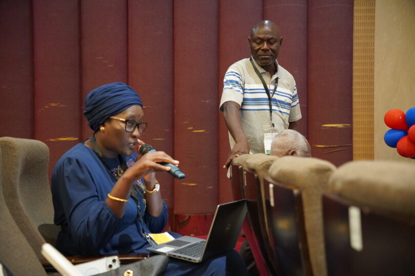 Eine Frau spricht in ein Mikrofon und hat dabei einen Laptop vor sich. Ein Mann steht neben den Sitzreihen und hört aufmerksam zu, während im Hintergrund rote, blaue und beige Elemente des Veranstaltungsraums zu sehen sind.