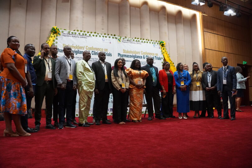 Mehrere Personen stehen in einer Reihe auf einer Bühne mit rotem Teppich vor einem Banner, auf dem die Stakeholders' Conference zu Pharmakovigilanz und klinischen Studien" angekündigt wird. Sie tragen Namensschilder und formelle Kleidung.