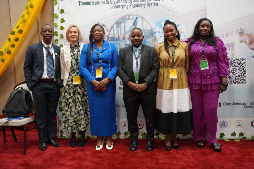 Eine Gruppe von sechs Personen mit Namensschildern und offizieller Kleidung posiert vor einem Banner auf dem zum Teil zu lesen ist: Theme: Medicine Safety Monitoring (...)"