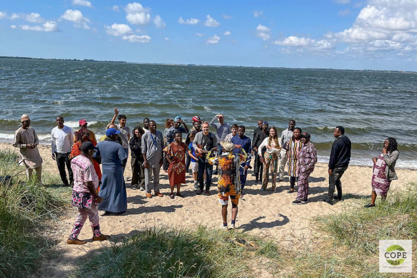 Eine Gruppe von Menschen steht am Strand mit Wasser, Sand und Düne und schaut auf eine Person, die vor der Gruppe steht.