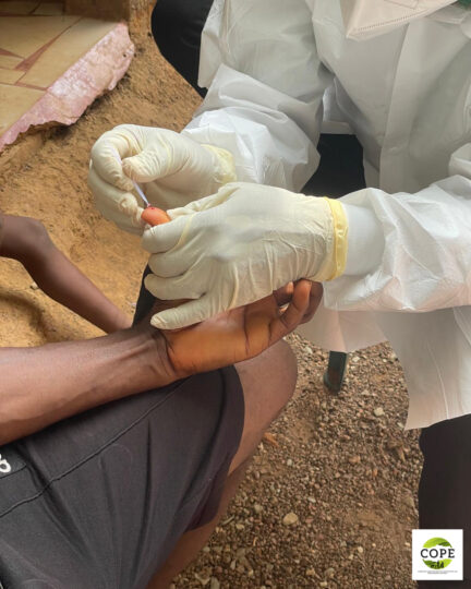 A seated person wearing a white coat, face mask, and white rubber gloves performs a finger-prick blood test on a second person sitting opposite.