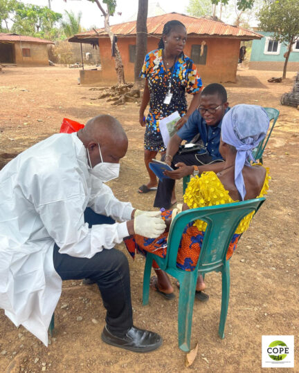 Outdoor: Four people at an open space in a village with reddish-brown gravel ground and mud huts with corrugated metal roofs. Three of the people are sitting together on plastic chairs. One of them is wearing a white coat, white rubber gloves, and a face mask, and is attending to the arm of the second person, who is wearing a blue headscarf. The third seated person is holding a small notebook and appears to be talking with the person in the blue headscarf.
