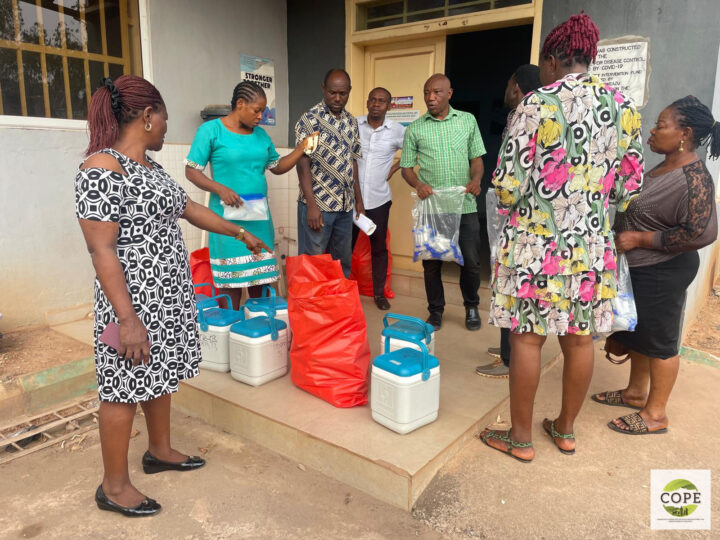 Outdoors: A group of people stands in front of a building entrance. They gathered around six plastic buckets with lids and handles, as well as several red bags and seem to discuss this equipment.