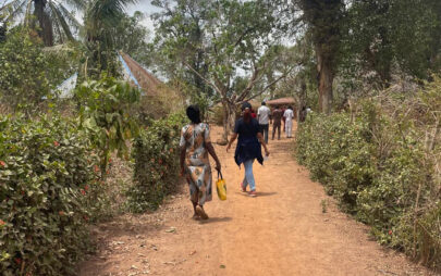 Eine Gruppe von Personen läuft einen Pfad aus rot-brauner Erde hinunter. Links und rechts des Pfades sind Büsche und Palmen.