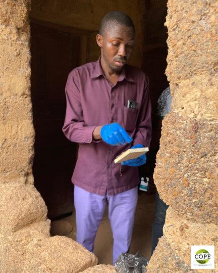 A man wearing blue rubber gloves standing on a sort of veranda. He is holding a small animal trap in his hands.