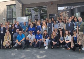 A group of people, smiling and posing for the photo , stands in front of the entrance of a building. In the back row of the group, there is a banner of the Robert Koch Institute.