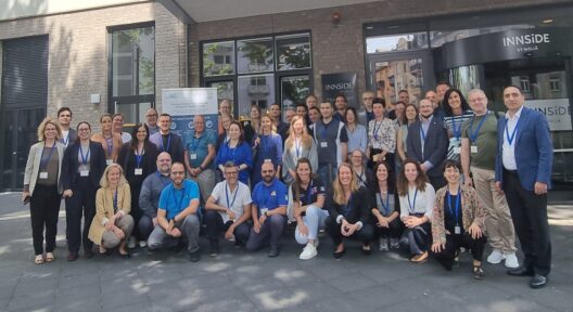 A group of people, smiling and posing for the photo , stands in front of the entrance of a building. In the back row of the group, there is a banner of the Robert Koch Institute.