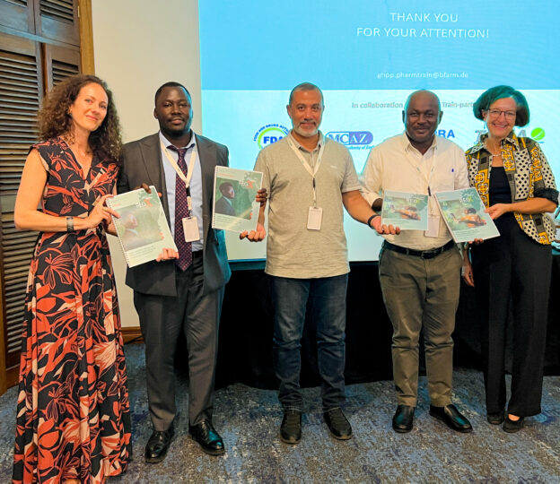 Group photo with five people holding A4 brochures and smiling at the camera. Behind them is a screened PowerPoint slide which says: ‘Thank you for your attention!ghpp.pharmtrain@bfarm.de.’