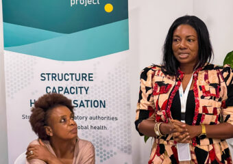 A talking woman stands next to a sitting woman who seemingly listens to her. In the background is a banner which says PharmTrain2 project.
