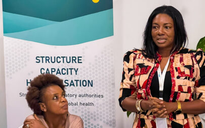 Eine Frau, die einen Konferenzausweis mit Band um den Hals trägt, steht redend neben einer zweiten, sitzenden Frau, die ihr zuzuhören scheint. Hinter der sitzenden Frau ist ein Banner mit unter anderem den Worten: "PharmTrain2 Project"