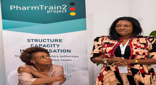 A talking woman stands next to a sitting woman who seemingly listens to her. In the background is a banner which says PharmTrain2 project.
