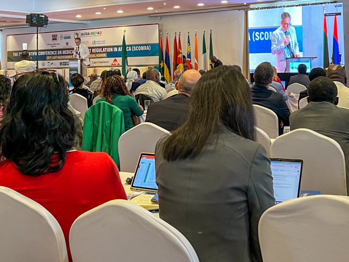 In a conference room with seating facing the stage. People, some with laptops, are sitting with their backs to the camera. On the stage, a man is standing behind a lectern, holding a microphone and speaking. Behind the man is a row of different national flags and a conference banner on the wall.