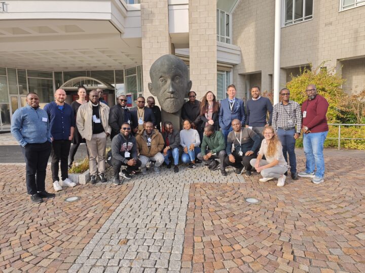 Eine mittelgroße Gruppe von Personen posiert vor dem Eingang des Paul-Ehrlich-Instituts (außen). Ein Teil der Gruppe steht links und rechts neben der Paul Ehrlich Büste und ein Teil der Gruppe sitzt in kniender Reihe davor. Viele haben Tagungsausweise um den Hals gehängt.