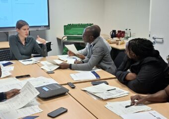 In a room with a large screen in the background, people are sitting at a seminar table in front of documents and notepads. One of the people is speaking and the others are listening.
