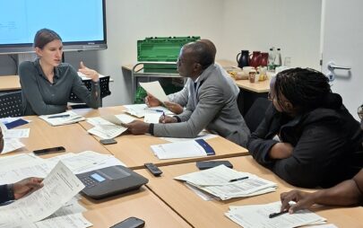 In a room with a large screen in the background, people are sitting at a seminar table in front of documents and notepads. One of the people is speaking and the others are listening.