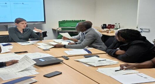 In einem Raum mit großem Bildschirm im Hintergrund sitzen an einem Seminartisch Personen vor Dokumenten und Schreibblöcken. Eine der Personen redet und die anderen hören ihr zu.