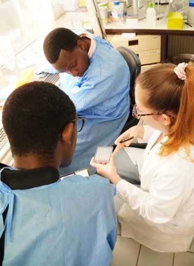 Three people in protective clothing are sitting in the laboratory and looking at a small pallet with samples.