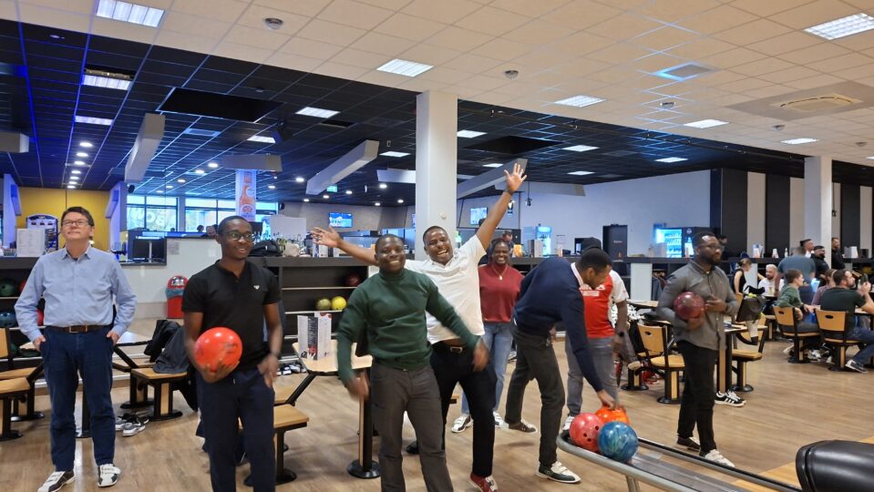 Innenraum einer Bowlingbahn. Ein paar Personen sind um eine Bowlingbahn versammelt und einige lachen fröhlich in die die Kamera.