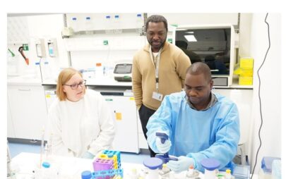 Laborszene: Eine Person mit Besucherausweis um den Hals hängend stehet hinter zwei sitzenden Personen, von denen einer an Laborgeräten tätig ist. Die zwei sitzenden Personen tragen blaue Schutzhandschuhe und Laborkittel.