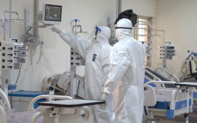 In einem Raum mit mit medizinischen Geräten ausgestatteten Krankenbetten stehen zwei Personen in Schutzanzügen, medizinischer Atemschutzmaske und Gesichtsschutz und schauen in einen medizinischen Monitor, der an der Wand hängt.