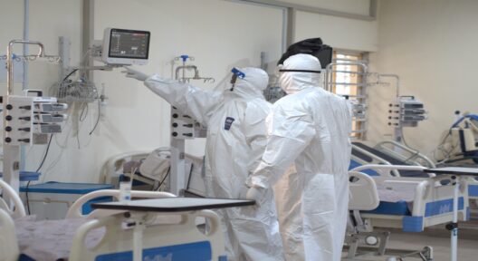 In einem Raum mit mit medizinischen Geräten ausgestatteten Krankenbetten stehen zwei Personen in Schutzanzügen, medizinischer Atemschutzmaske und Gesichtsschutz und schauen in einen medizinischen Monitor, der an der Wand hängt.