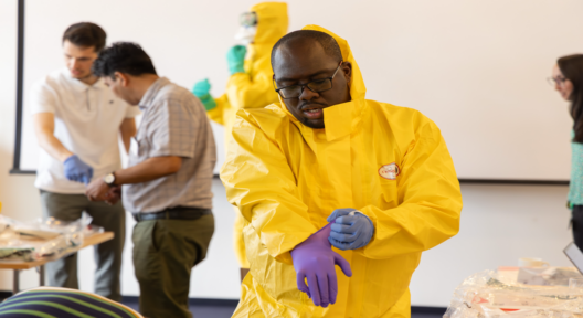 A man in a yellow protective suit is putting on medical rubber gloves