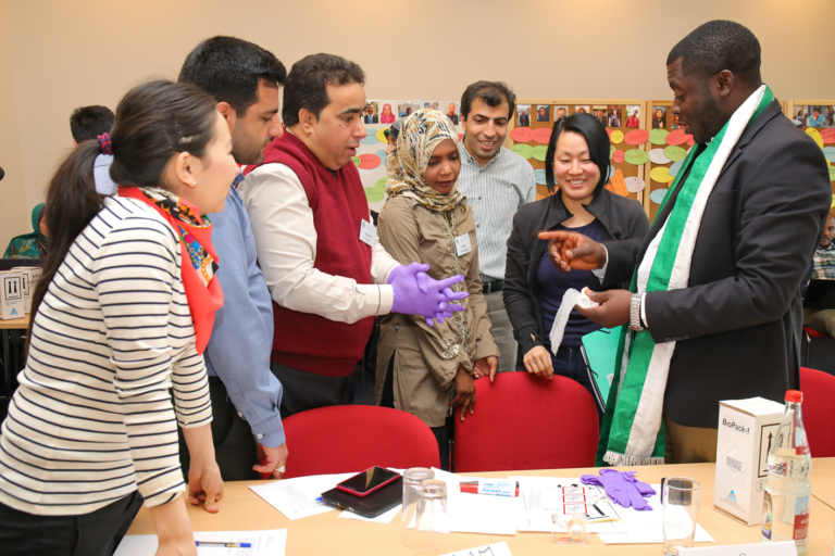 Eine Gruppe von Menschen, von denen einige Namensschilder tragen, steht in einem Workshop- oder Seminarraum und unterhält sich miteinander.