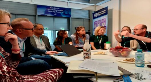 Gruppe von Personen mit Namensschildern um den Hals tragend und am Tisch sitzend. Eine der Personen redet und gestikuliert.