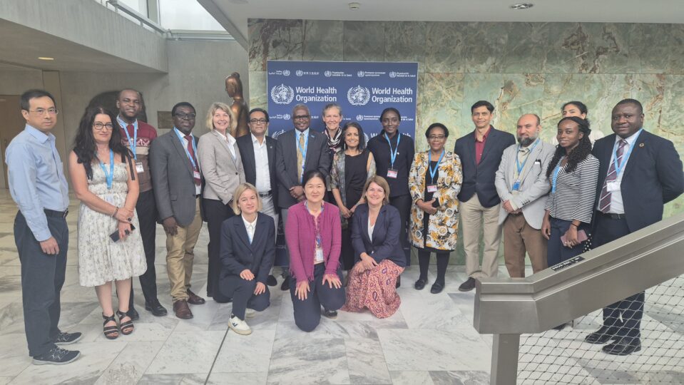 Eine Gruppe von Personen posiert für das Foto vor einem Banner mit Aufschrift "World Health Organzation" in einem Flur mit Mamorwand und Mamorfussboden eines Gebäudes